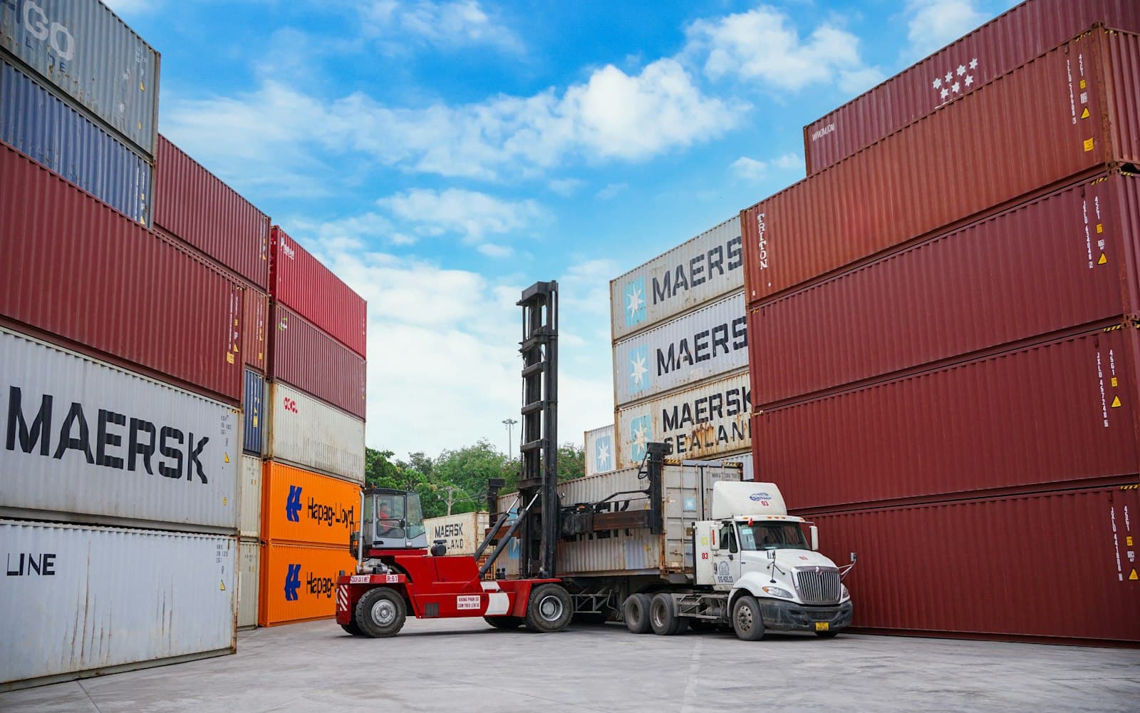 Stock photo of a container yard with stacked shipping containers, a reach stacker and a truck